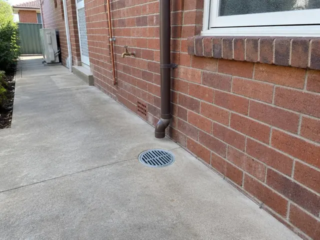 Outdoor gully grate on dry concrete beside a brick house wall and downpipe in Arncliffe