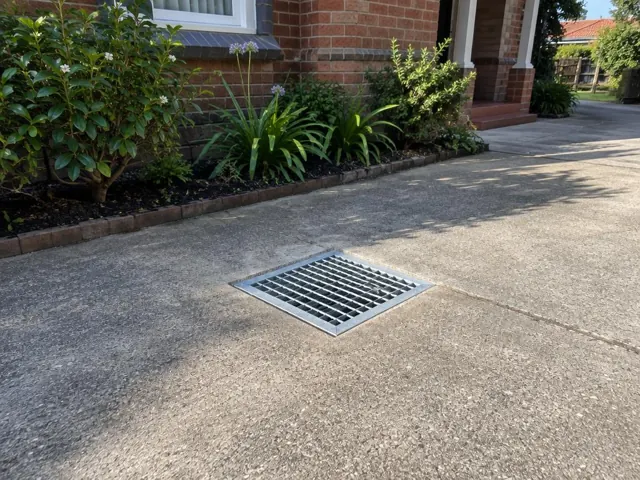 Stormwater grate seated in a concrete driveway beside a brick house in Bexley