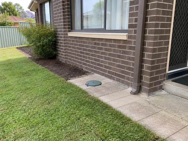 Capped sewer access cover beside brick house wall and paved path in Kingsgrove